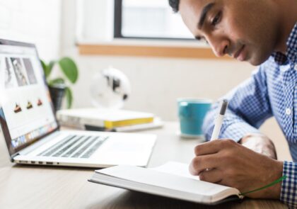 kuvituskuva, jossa mies pöydän ääressä kirjoittaa muistikirjaan edessään kannettava tietokone