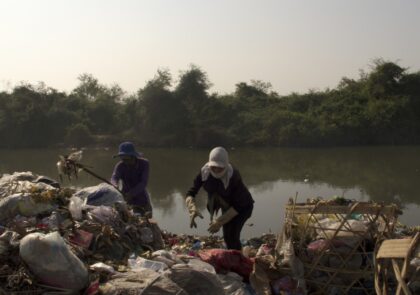 Valokuvassa taustalla joki ja etualalla mies ja nainen, jotka tutkivat joenpenkalla olevaa jätekasaa.