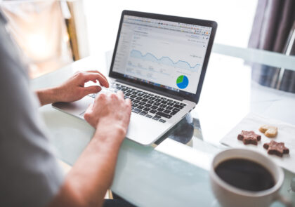 Kuvassa on kannettavalla tietokoneella kirjoittava ihminen. Kuvassa on myös kuppi kahvia ja kolme pikkuleipää.