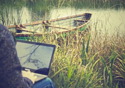 kuvituskuvana valokuva, jossa henkilö istuu rannalla kannettava tietokone sylissään. Taustalla näkyy vettä ja soutuvene.