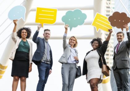 artikkelikuvana valokuva, jossa joukko hymyileviä ihmisiä pitää päänsä päällä erivärisiä tyhjiä puhekuplia.