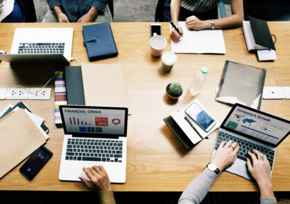 Kuvassa on neljän toisiaan vastapäätä istuvan ihmisen kädet. Kaksi työskentelee laptopeilla, yksi kirjoittaa paperille ja yhden kädet ovat sylissä. Pöydällä on vesipullo ja kaksi kahvimukia sekä pieni huonekasvi.