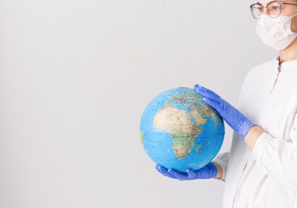 artikkelikuvassa kasvomaskiin ja sinisiin hanskoihin pukeutunut nainen pitelee käsissään maapalloa.