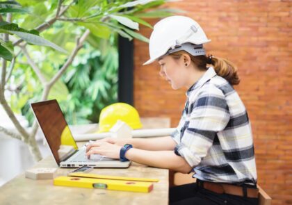 Tietokoneella työskentelevä nainen, jolla päällään ruutupaita ja valkoinen turvakypärä.