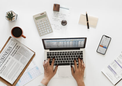 Kuvituskuvassa ihmisen kädet näpyttelevät tietokoneen näppäimistöä ja pöydällä on mysö laskin, paperia ja kynä sekä kahvikuppi.