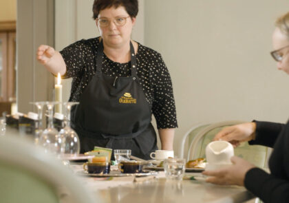 Kaksi naista kahvipöydän ääressä, toinen sytyttää kynttilää.