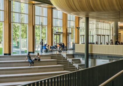Valokuva puurakenteiselta kampukselta, etualan portailla istuu opiskelijoita ja taustalla on suuret ikkunat.