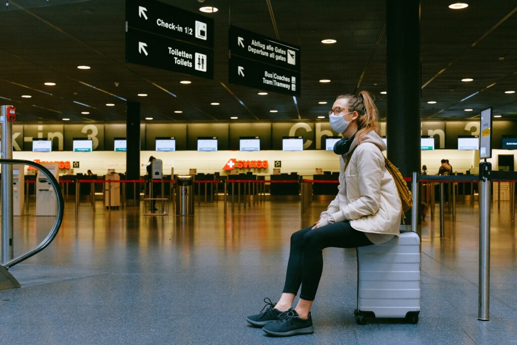 A person with a mask sitting on a suitcase at the airport.