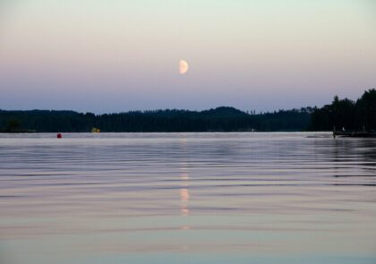 Kesäöinen järvimaisema, jossa kuu heijastuu vedenpinnasta.