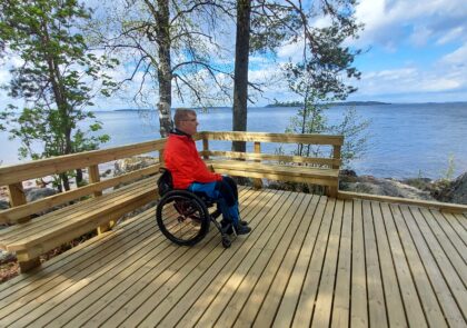 Kuvituskuvassa ulkoilija istuu pyörätuolissa isolla puisella terassilla veden äärellä.