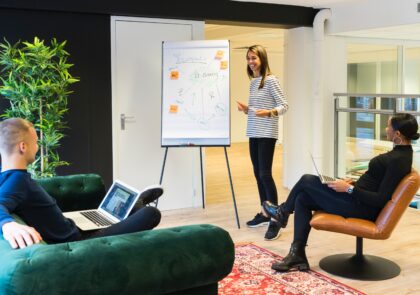 Ihmisiä nojatuoleilla jotka katsovat fläppitaulun vieressä seisovaa naista