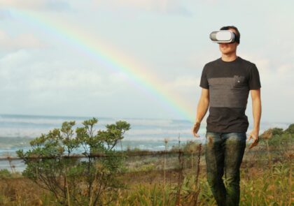 Mies kävelee virtuaalimaailmassa rannalla 3D-lasit päässään, taustalla sateenkaari.