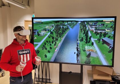 Kuvassa henkilö seisoo huoneen keskellä virtuaalitodellisuuslasit päässään ja ohjain molemmissa käsissä. Henkilön kädet ovat kohotettuna rinnan korkeudelle ja hänen takanaan on suuri televisioruutu. Television ruudulla näkyy virtuaalisen harjoitteluympäristö.