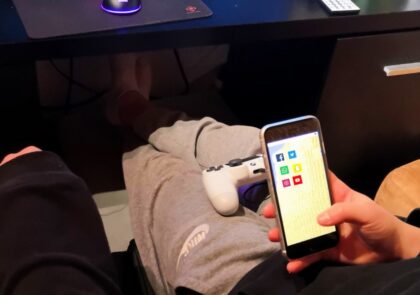 Kuvituskuvassa ihminen istuu ja näpyttää kännykkää. Sylissään hänellä on myös pelikonsolin ohjain.