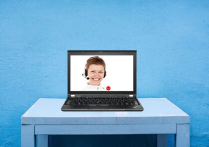 Kuvituskuvassa tietokone pöydällä ja tietokoneen ruudulla naisen kasvot ja kuulokkeet korvissa.