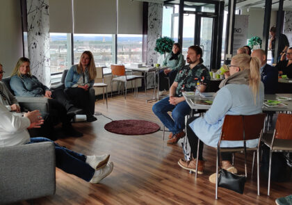 Joukko ihmisiä istuu pöytien ääressä ja seuraa ikkunoiden edessä tapahtuvaa paneelikeskustelua