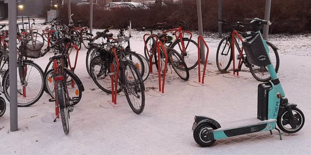 Yhteiskäyttösähköpotkulauta ja polkupyörät pyöräkatoksessa lumisessa maisemassa.