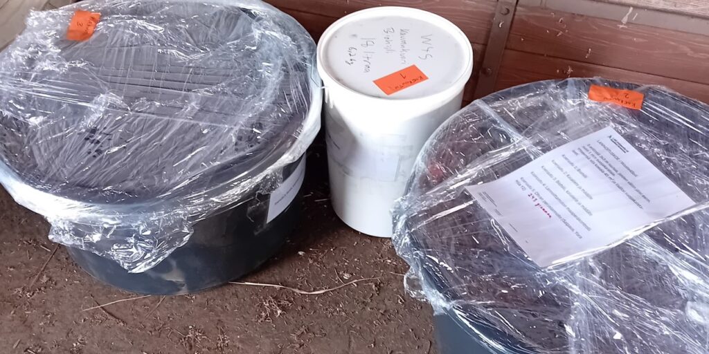 Kaksi mustaa saavia on peitetty kiristyskalvolla. Saavien vieressä on valkoinen säilytysastia, jossa on kansi päällä