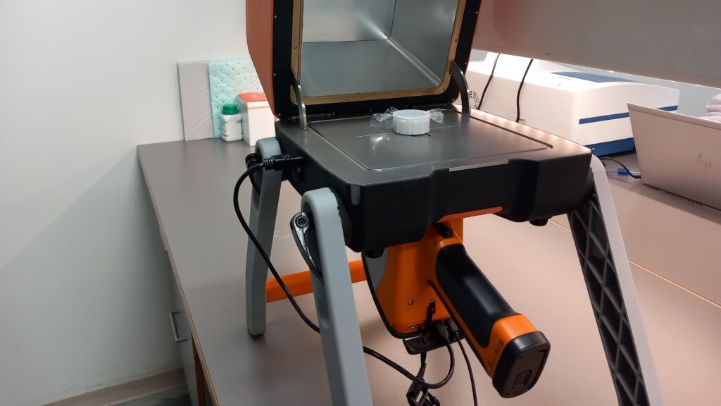 Oranssin säteilysuojan alle on kiinnitetty analysaattori, jossa on kahva, mutta näyttö ei näy tästä kulmasta. Säteilysuojassa on yksi näytekuppi.