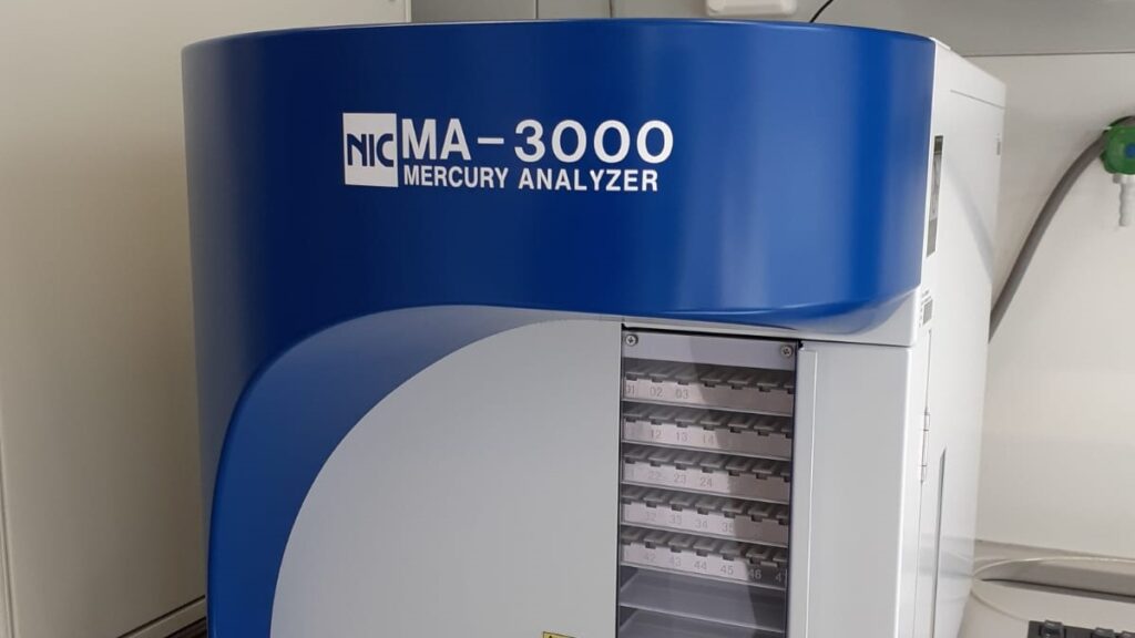 Neliskanttinen analysaattori on pöydällä ja väriltään harmaa ja sininen. Laitteen yläreunassa lukee NIC MA-3000 Mercury analyzer. Laitteen oikeassa reunassa on ikkuna, josta näkyvät hyllyillä olevat näytetelineet.