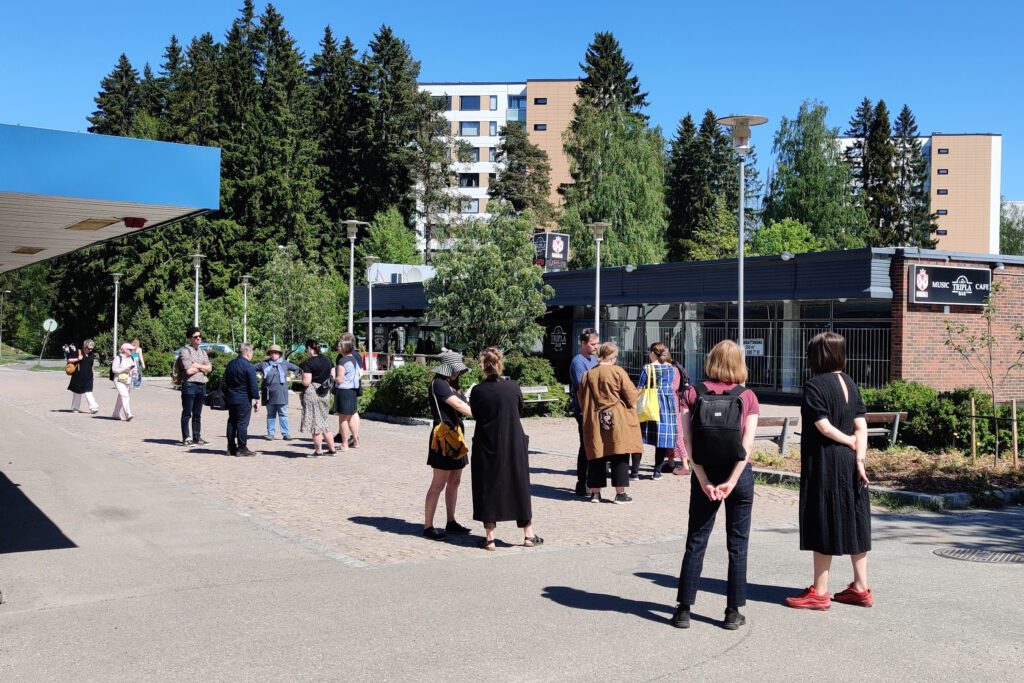 Joukko ihmisiä seisomassa kahden rakennuksen välissä aukiolla. Taustalla korkeita kuusia ja kerrostaloja. 