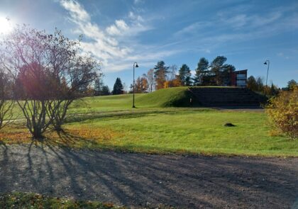 Nurmikkopeitteinen puisto, jossa kumpare, muutama puu, katulamppu sekä etualalla kävelytie. Horisontissa kerrostaloja ja puita.