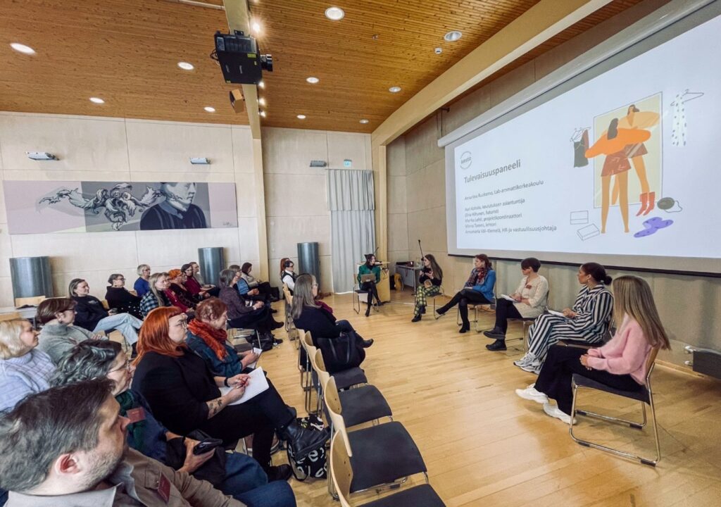 Seminaaritila, jonka edessä kuusi ihmistä istuu rinnakkain paneelikeskustelussa yleisön katsoessa. Taustalle videotykillä heijastettuna otsikko Tulevaisuuspaneeli.