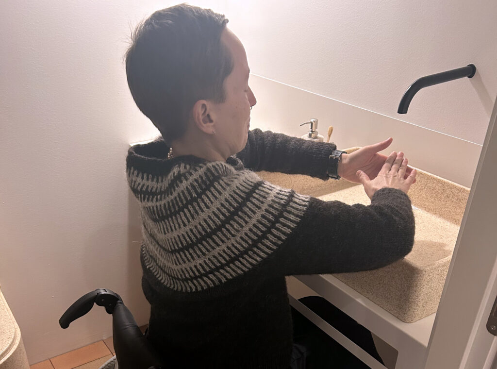 Valokuva, jossa pyörätuolissa istuva henkilö pesee käsiään korkealla olevan käsienpesualtaan ääressä.