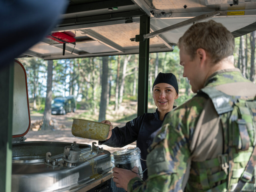 Etualalla armeijan vaatteissa mies selin kameraan, hänen edessään hymyilevä nainen ojentaa soppatykistä peltisellä astialla ruokaa. Taustalla metsää.