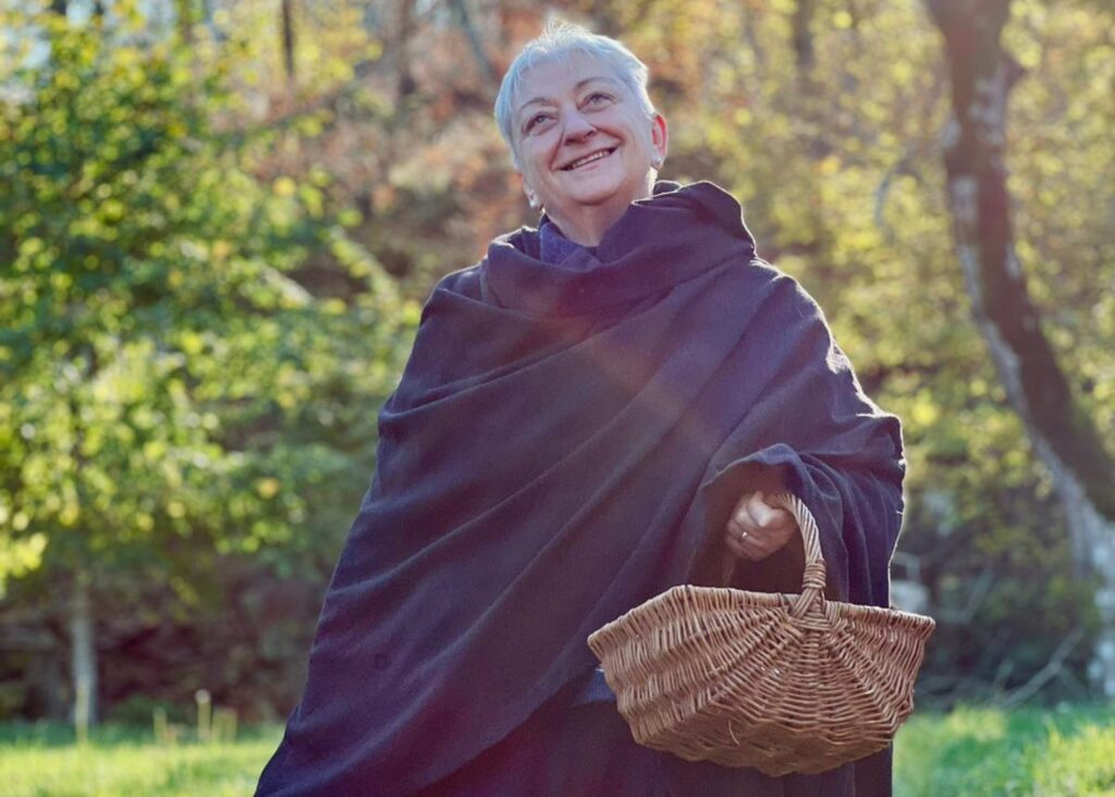 An elderly woman standing, holding a basket. Forest in the background.