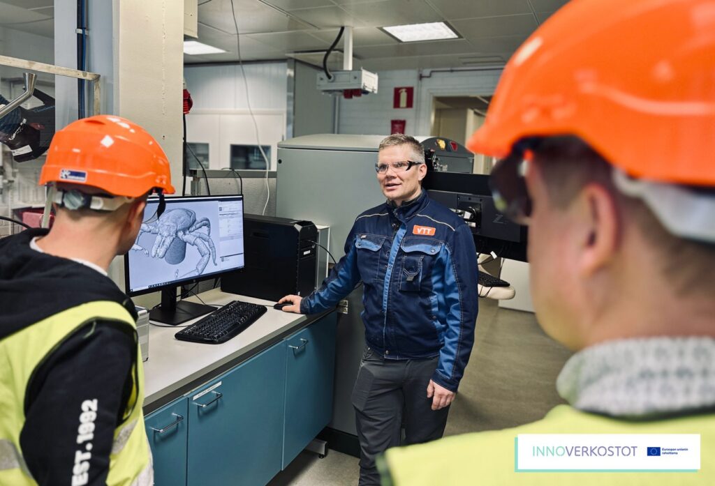 Kolme henkilöä, joista kaksi oransseissa turvapäähineissä seuraamassa kolmannen pitämää VTT Jyväskylän mikrotomografia-kuvantamislaitteiston esittelyä.