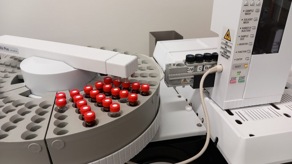 An analysis device with small sample bottles on the sample tray. The robot arm is ready to transfer the bottles to the carriage of the injector unit, where the washing bottles are ready and waiting.