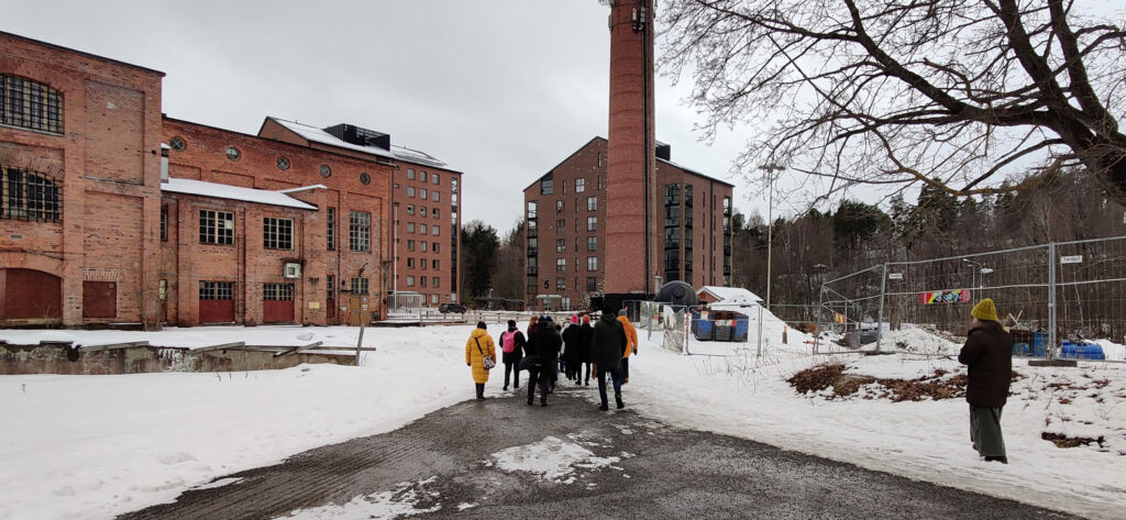 Ryhmä ihmisiä kuuntelee opasta punatiilisen rakennuksen (Kankaan vanha sauna) edustalla.