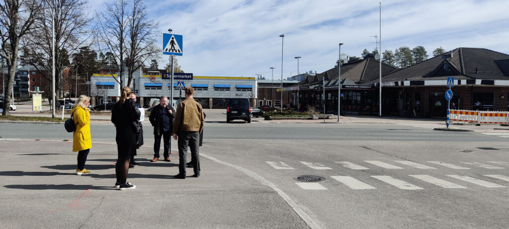 Ryhmä ihmisiä seisoo kadun kulmassa suojatienvieressä. Taustalla matalia tummakattoisia rakennuksia ja K-kauppa.