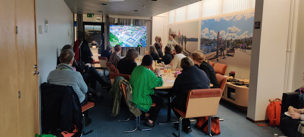 Ryhmä ihmisiä istuu pöydän ääressä selin kameraan ja katsoo näytöltä esitystä.