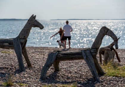 Etualalla puusta veistettyjä hevosia ja taustalla kaksi ihmistä leikittää koiraa Saimaan rannalla.
