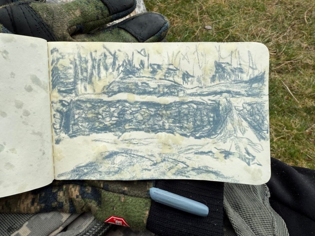 Lähikuvassa ulkotilassa rukkaseen puettu käsi pitelee luonnoslehtiötä, johon on piirretty harmaalla vahaliidulla luonnosta kivimuurista. 