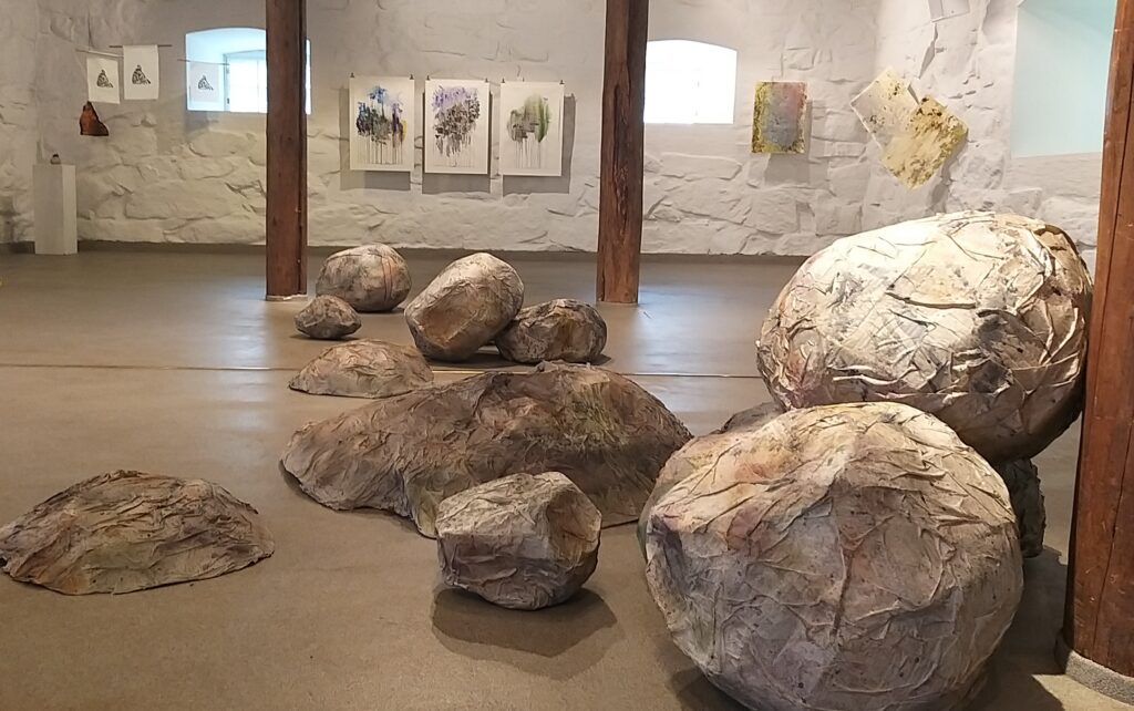 Kuvassa on näyttelytila, jossa on beigenvärinen muovinen lattia, ruskeat puiset pylväät ja valkoiseksi kalkitut kivinavetan seinät. Kuvassa etualalla näkyy tilaan sijoitettuna suuria kiviä muistuttavia lohkareita. Taustalla näkyy maalauksia ja grafiikkaa.