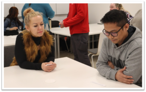 Kaksi ihmistä istuu pöydän ääressä. Taustalla näkyy muita ihmisiä.