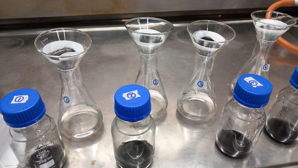 Säilöpulloja, joissa on mustaa lientä pohjalla ja siniset korkit, joihin näytteen numero on merkitty. Kirkasta nestettä on suodatettu numeroituihin Erlenmeyer-pulloihin, joissa on lasisuppilot sekä taitetut suodatinpaperit, joissa on mustaa kiintoainesta.]