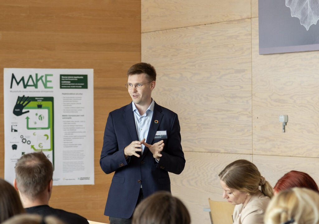 Timo Huhtamäki, nuori mieshenkilö silmälaseissa ja sinisessä puvuntakissa, seisoo yleisön edessä puhumassa. Taustalla MAKE-hankkeen juliste.