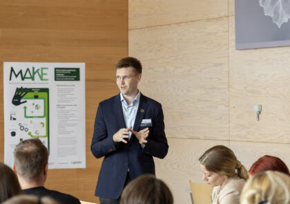 Timo Huhtamäki, nuori mieshenkilö silmälaseissa ja sinisessä puvuntakissa, seisoo yleisön edessä puhumassa. Taustalla MAKE-hankkeen juliste.