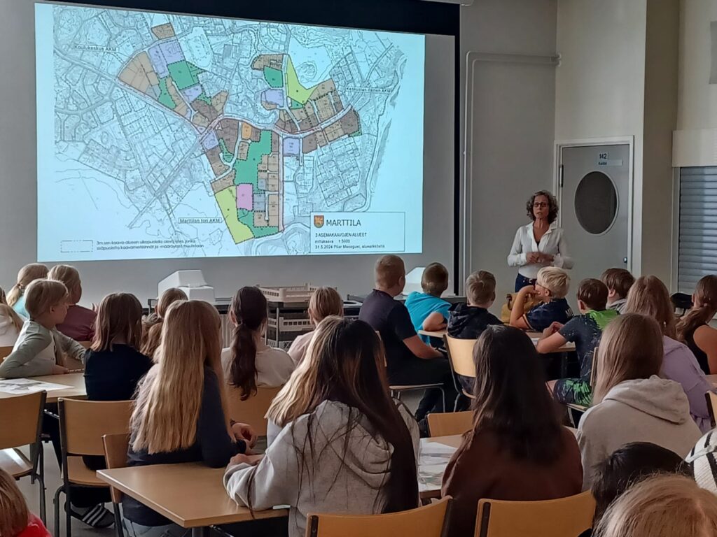 Valokuvassa asiantuntija esittelee koululaisille seinälle heijastettua kuvaa asemakaavakartasta.