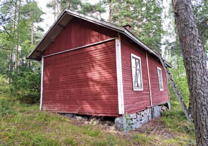 Kuva esittää pientä hirsirunkoista mökkiä, joka on väritykseltään punainen, nurkkalaudat valkoiset. Rakennuksessa on luonnonkiviperustukset.