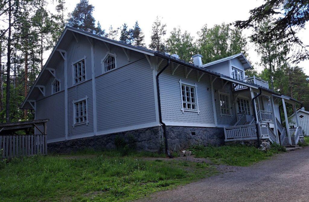 Kuva esittää suurta hirrestä rakennettua asuinrakennusta, joka on väritykseltään vaalea ja vuorilaudoitus on valkea. Rakennuksessa on runsaasti koristeellisia yksityiskohtia.